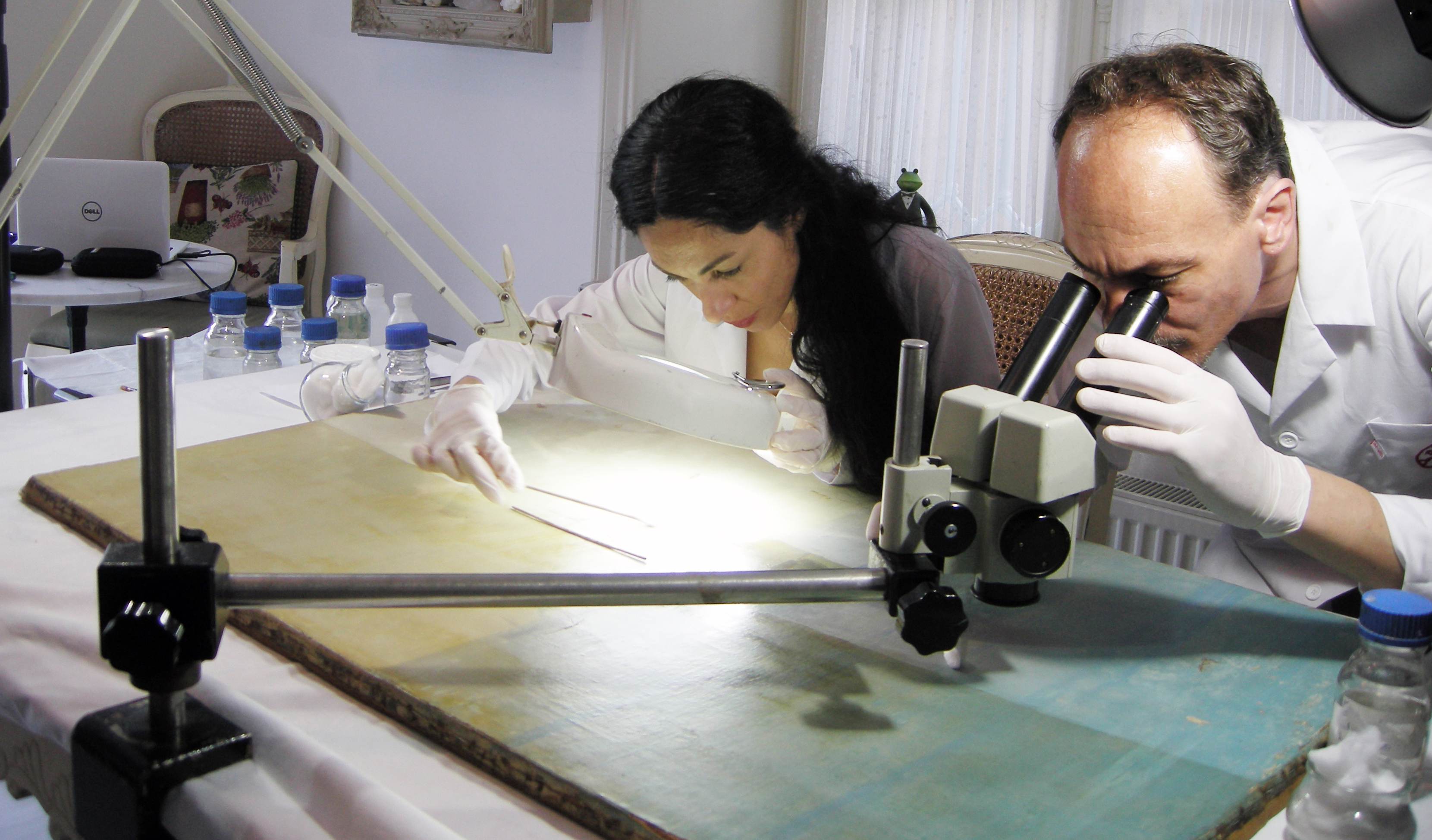 Two conservators examine the surface of a panel painting under raking light, one inspecting through a stereo microscope.